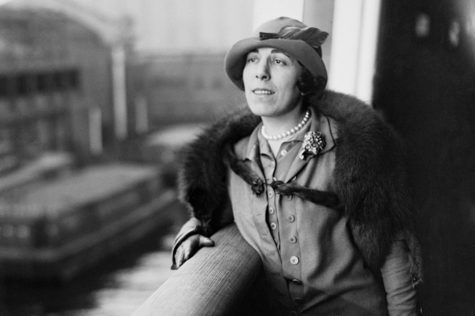 Photograph of Edna Ferber, wearing a hat, pearls, fur stole, suit, and gloves, leaning on a railing overlooking a body of water.