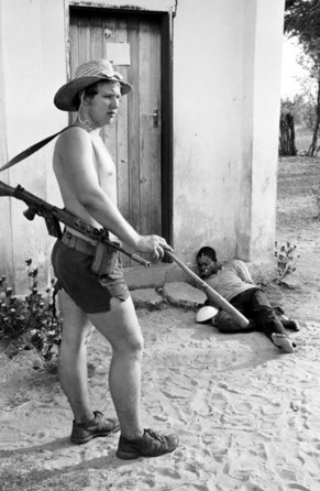 Lt. Graham Baillie raps a small wooden bat against his leg after using it to beat Moffat Ncube, a local teacher, political leader and now a bound, unconscious prisoner slumped against the wall of school house, 20 September 1977. Ncube reportedly later died after three days of brutal, non-stop torture.
