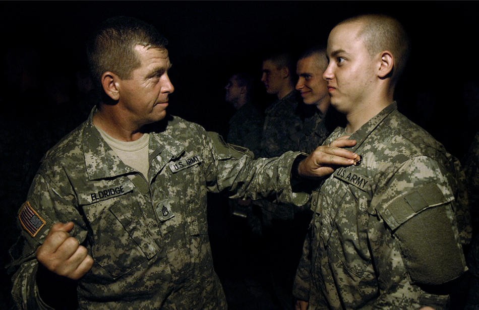 Fisher braces for his crossed-rifles pin, signifying his completion of training.  Drill sergeant John Eldridge holds the pin in place and prepares to secure it with a fist. "I hit him square on the chest," Eldridge said. "He had two holes, and blood dripping down from each - 'blood rifles.'"  Thirteen weeks of basic training had led Fisher to this—a 10-mile march through a series of high-stress combat tests that ended at a place called Honor Hill.  A sergeant closed the ceremony Sept. 17